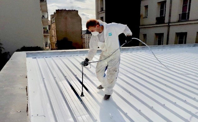 Un applicateur en combinaison blanche pulvérise le revêtement réfléchissant sur un toit en ville.